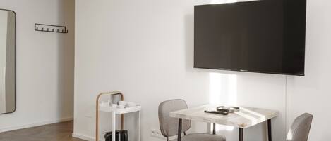 Un salón moderno con una mesa blanca, dos sillas grises, un televisor montado en la pared y un carro blanco con utensilios de cocina.