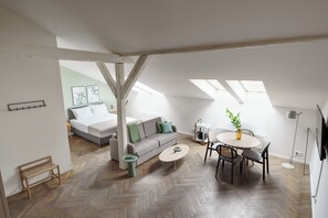 Un loft espacioso con piso de madera en patrón de espiga, un sofá gris con cojines verdes, una mesa redonda con sillas y un pequeño banco.