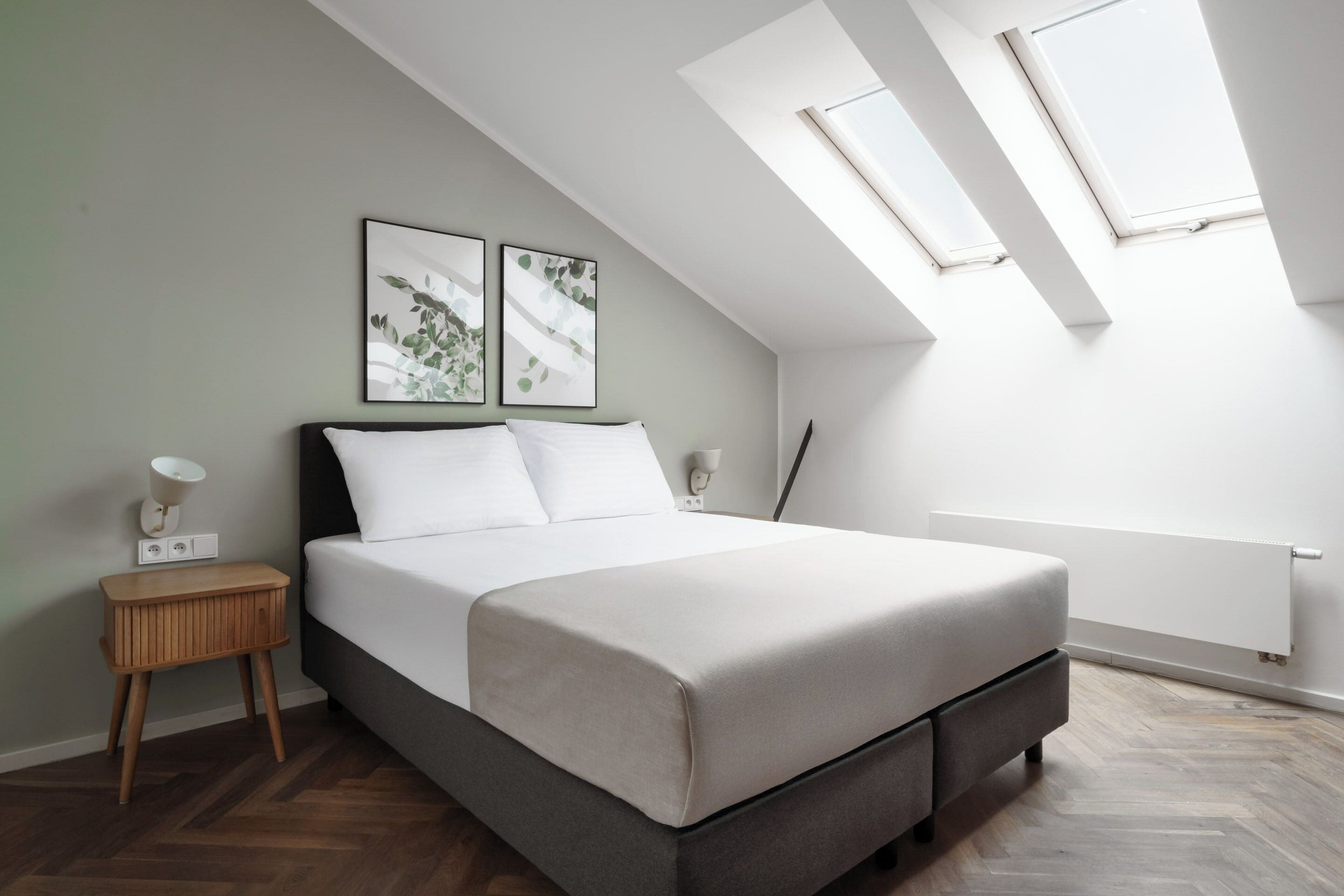 A modern bedroom with a bed, a nightstand, a wall-mounted lamp, and two skylights.