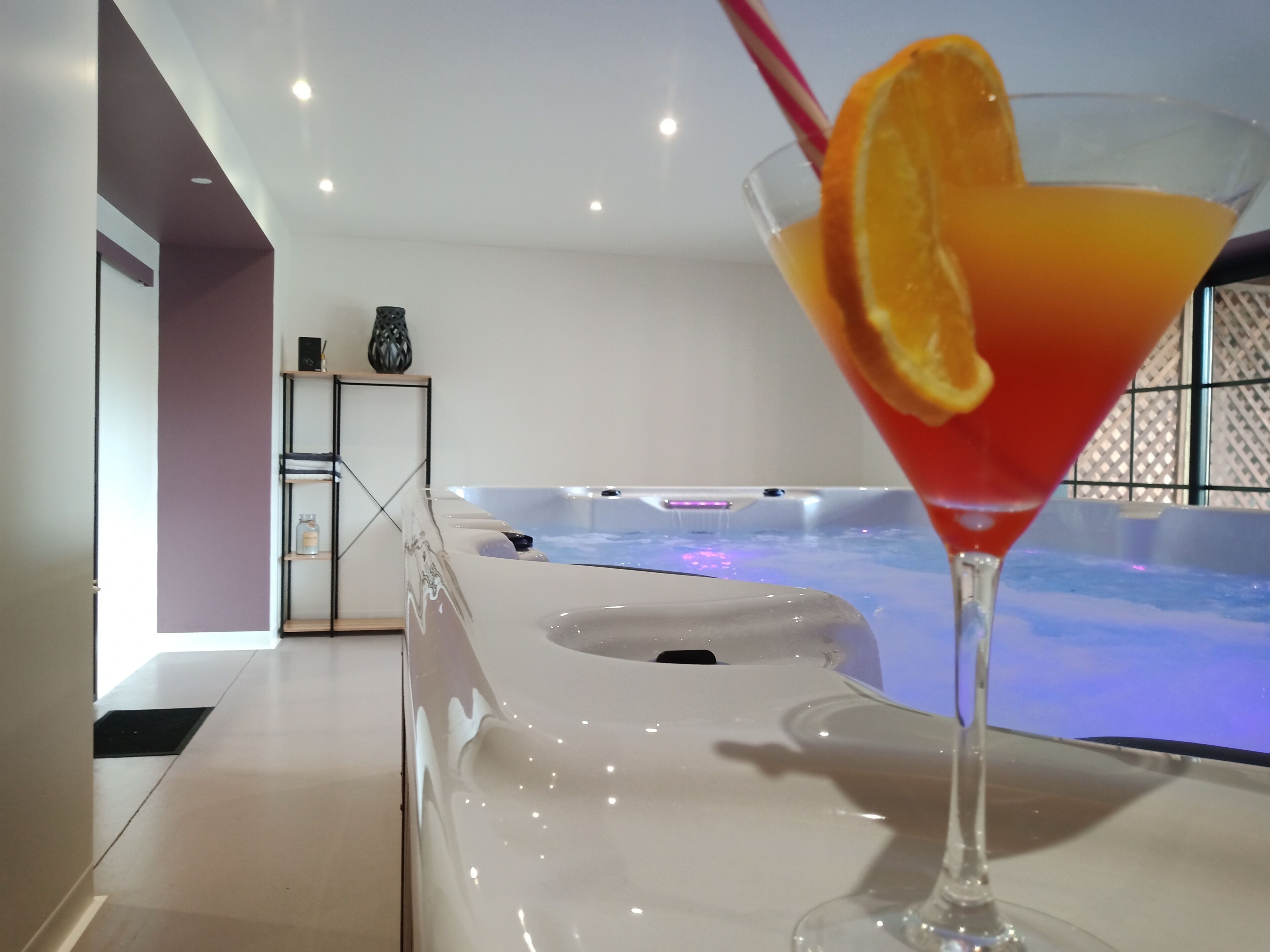 A modern interior with a glass table, a martini glass with an orange garnish, and a hot tub in the background.
