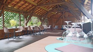An outdoor dining area with wooden tables and chairs, a covered ceiling, and a view of greenery through a lattice. - L'Hacienda (Tartas)