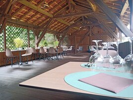 Un área de comedor al aire libre con mesas y sillas de madera, techo cubierto y vista a la vegetación a través de una celosía.