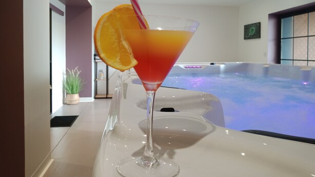 A martini glass with an orange slice and a pink straw on a table, with a hot tub in the background. - L'Hacienda (Tartas)