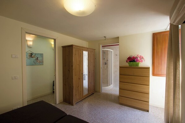 A room with a bed, a wooden wardrobe, a bathroom with a shower, and a dresser with a vase of flowers.