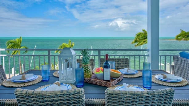 A dining table set on a balcony with a view of the ocean, including a bottle of wine, fruit, and blue glassware. - World-Class 5 Bed Villa  Oceanfront Pool/Hot tub Sunset Escape Sleeps 12 (Marathon)