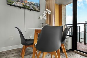 A modern dining area with a table and two chairs, a vase with white flowers, a television on the wall, and a view of the city through a window.