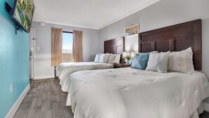 Una habitació d'hotel amb dos llits, una finestra gran amb cortines, un capçal de llit de fusta i una televisió penjada a la paret.