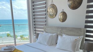 Una cama grande con ropa de cama blanca, un plaid amarillo estampado y toallas dobladas con cuidado. Hay dos platos decorativos colgados en la pared y una alfombra de cuadros sobre el suelo de madera.