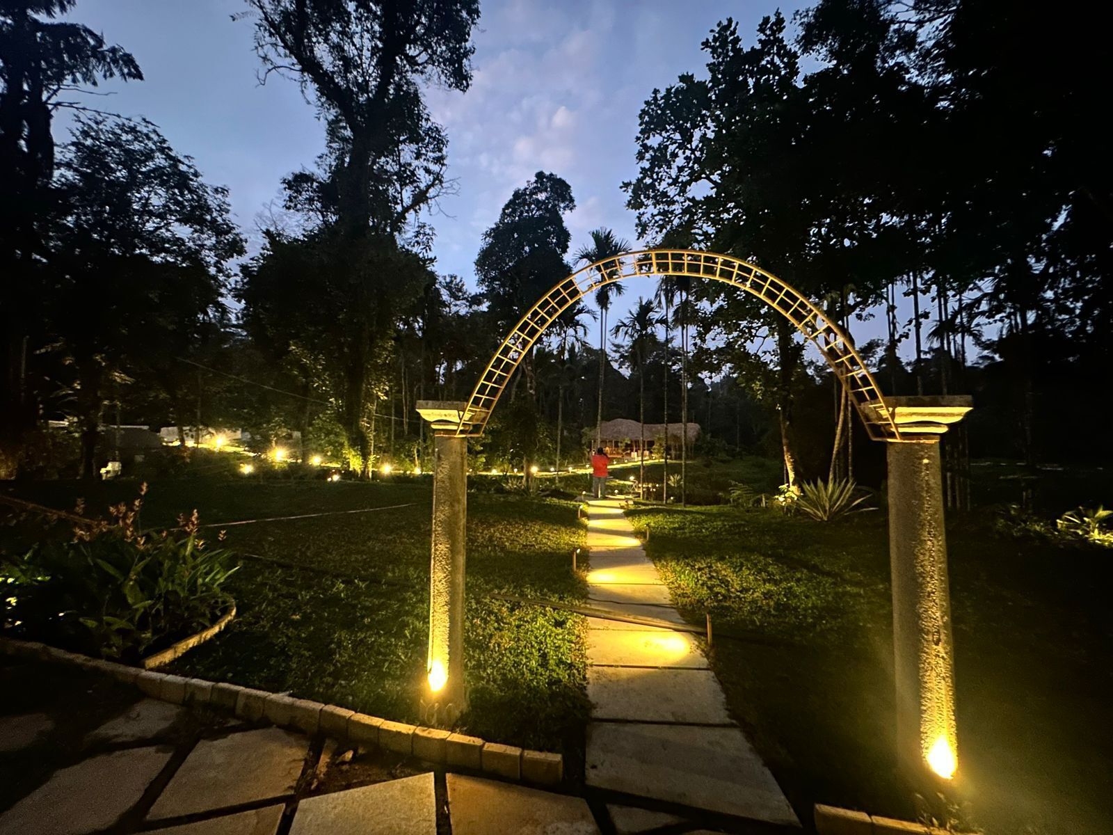 Un sendero conduce a una estructura de techo curvo, rodeada de árboles e iluminada por una cálida luz.