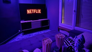 Uma sala de estar acolhedora com uma televisão de tela plana exibindo o logotipo da Netflix, um sofá macio, potes de pipoca e uma janela com persianas.