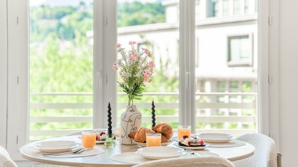 Mesa de café da manhã com pratos, talheres, um vaso com flores, croissants e copos de suco.