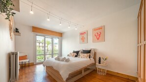 Quarto com uma cama grande, uma cadeira, um radiador, uma mesinha redonda e duas fotos emolduradas na parede.