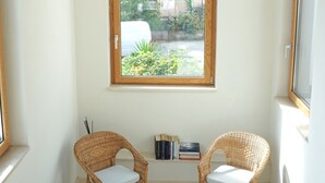 A corner room with two wicker chairs, a small shelf with books, and a window with a view of greenery and buildings. - La Ginestra - Antheia Homes (Scafati)