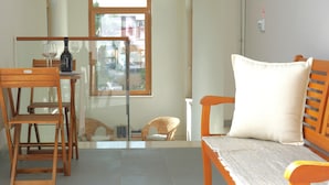 Une petite pièce bien éclairée, avec une table en bois, deux chaises, un canapé avec un coussin et une porte vitrée donnant sur un balcon.