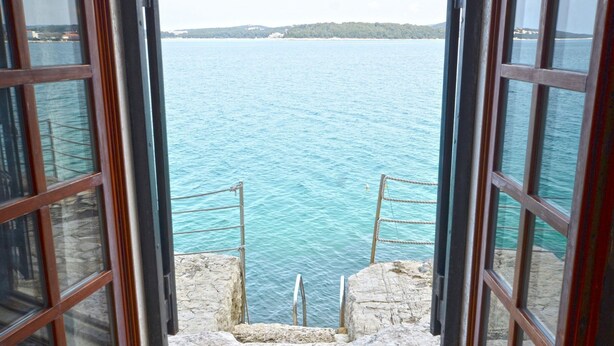 Vista a través de una ventana que muestra una escalera de piedra que lleva a un balcón con vista al agua.