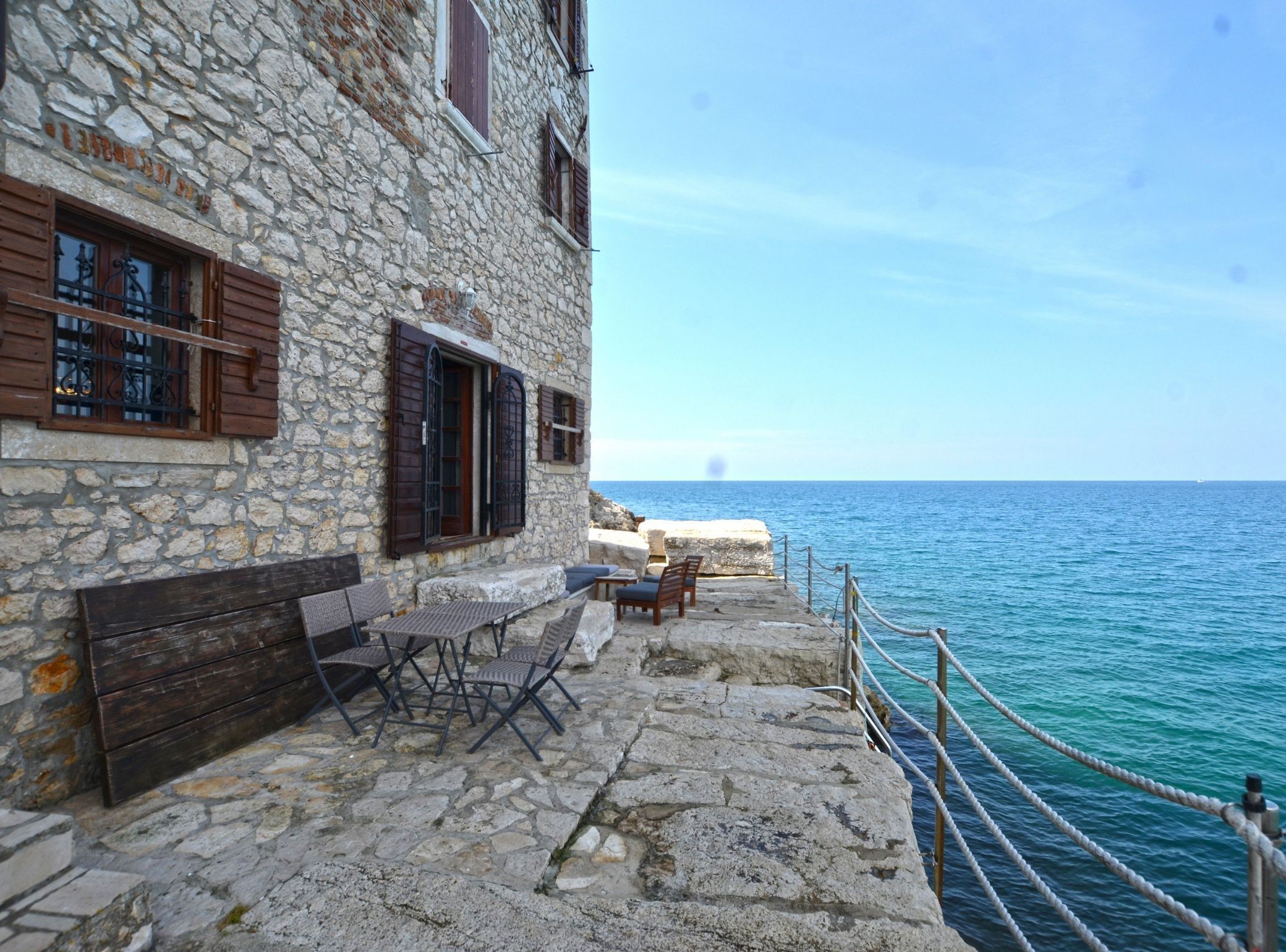 Ein Steinhaus mit einer gepflasterten Terrasse, Holzbänken und Tischen mit Blick auf das Meer.
