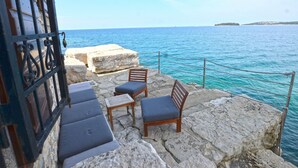 Un patio de piedra con mobiliario exterior frente al mar, que incluye una mesa y dos sillas.