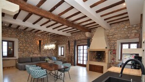 Amplia sala de estar con pared de piedra, chimenea, mesa de comedor con sillas y zona de cocina con fregadero y estufa.