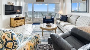 A living room with a TV, sofa, armchair, and a view of the beach through the sliding glass doors. - Beach House 201D - Gulf Front Resort! (Miramar Beach)