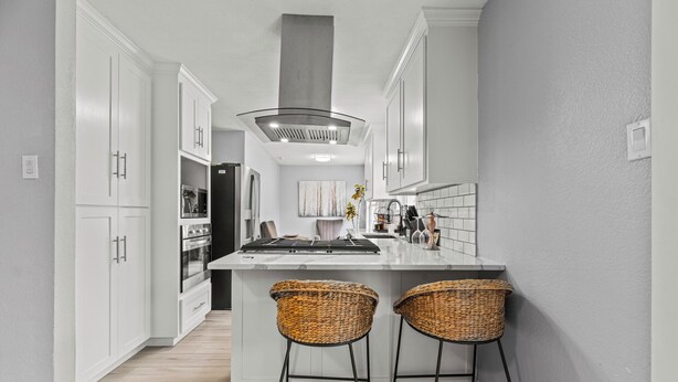 A modern kitchen with white cabinetry, a central island with bar stools, and a stainless steel range hood. - Sunset Heights Retreat- 3 Stylish & Spacious Bedroom (Fort Worth)