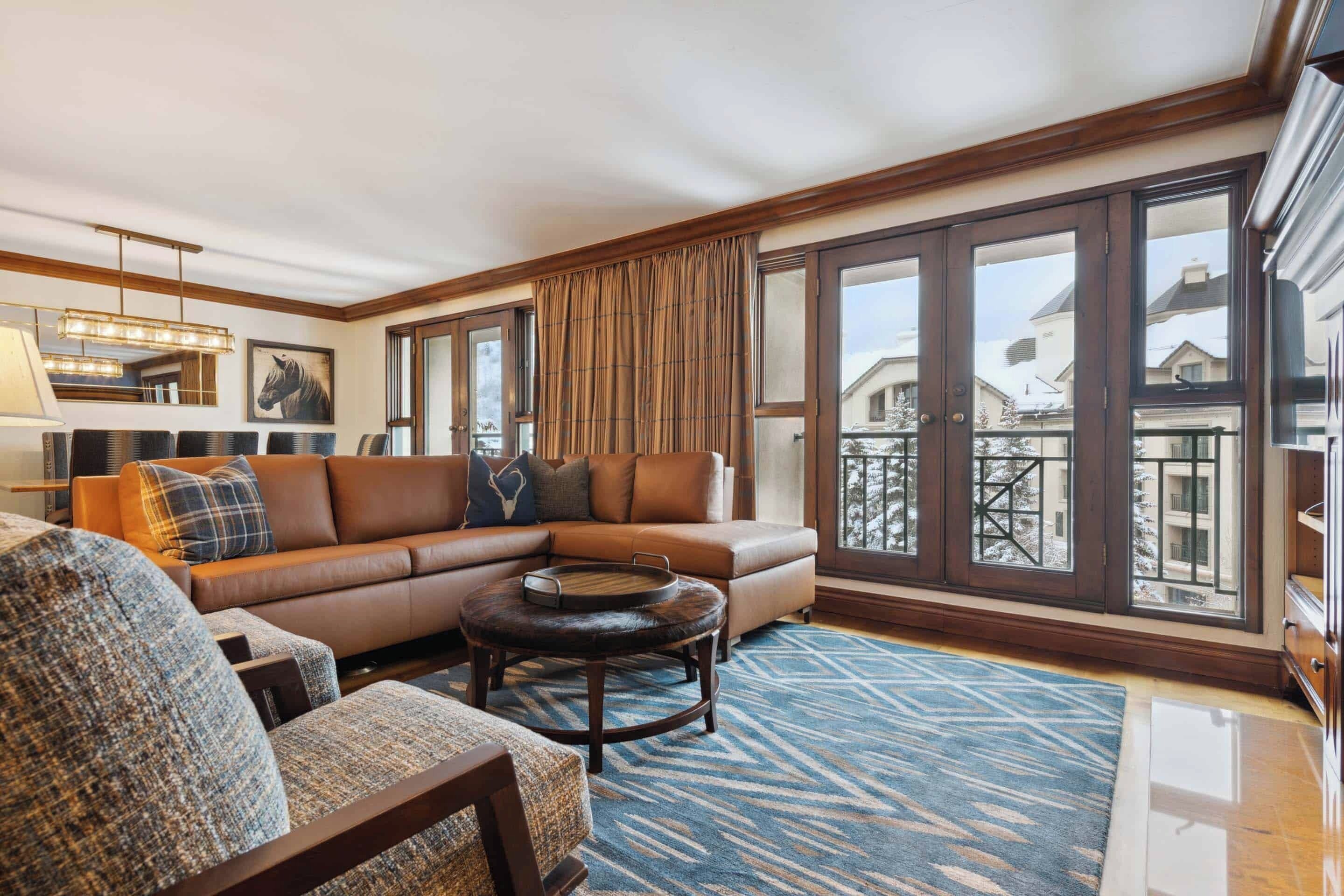 Un salon moderne avec un canapé en cuir marron, un fauteuil à motifs et une table basse ronde. De grandes fenêtres offrent une vue sur l’extérieur.