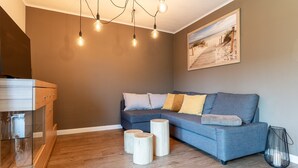 A modern living room with a grey sofa, wooden side tables, and a painting of a beach scene. - Apartment in Busum Near Perlebucht Lagoon (Westerdeichstrich)