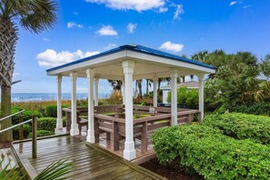 A covered outdoor area with a wooden deck and benches, surrounded by greenery and a view of the ocean.