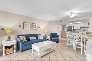 A living room with a blue sofa, white coffee table, and a dining area with white chairs and a wooden table. - Luxury Ocean View Condo- Relaxing Exclusive Access (Myrtle Beach)