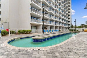 A hotel with a swimming pool, multiple balconies, and a paved walkway.