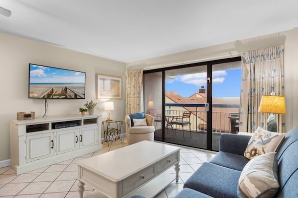 A living room with a sofa, a coffee table, a TV, a balcony with a view of buildings, and a sliding door.