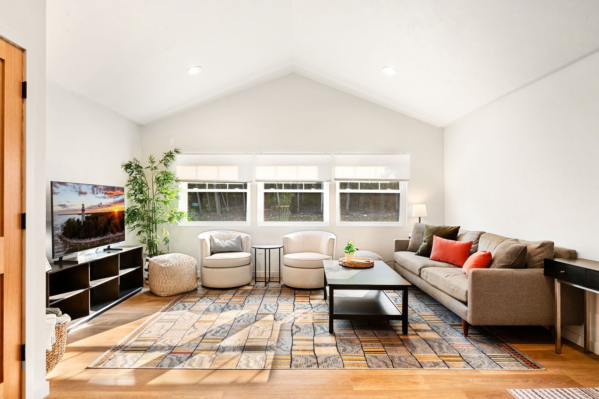 A modern living room with a high ceiling, a large window, a flat-screen TV, a sofa, two armchairs, a coffee table, and a patterned rug.