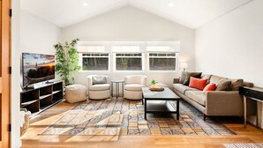 A modern living room with a high ceiling, a large window, a flat-screen TV, a sofa, two armchairs, a coffee table, and a patterned rug.
