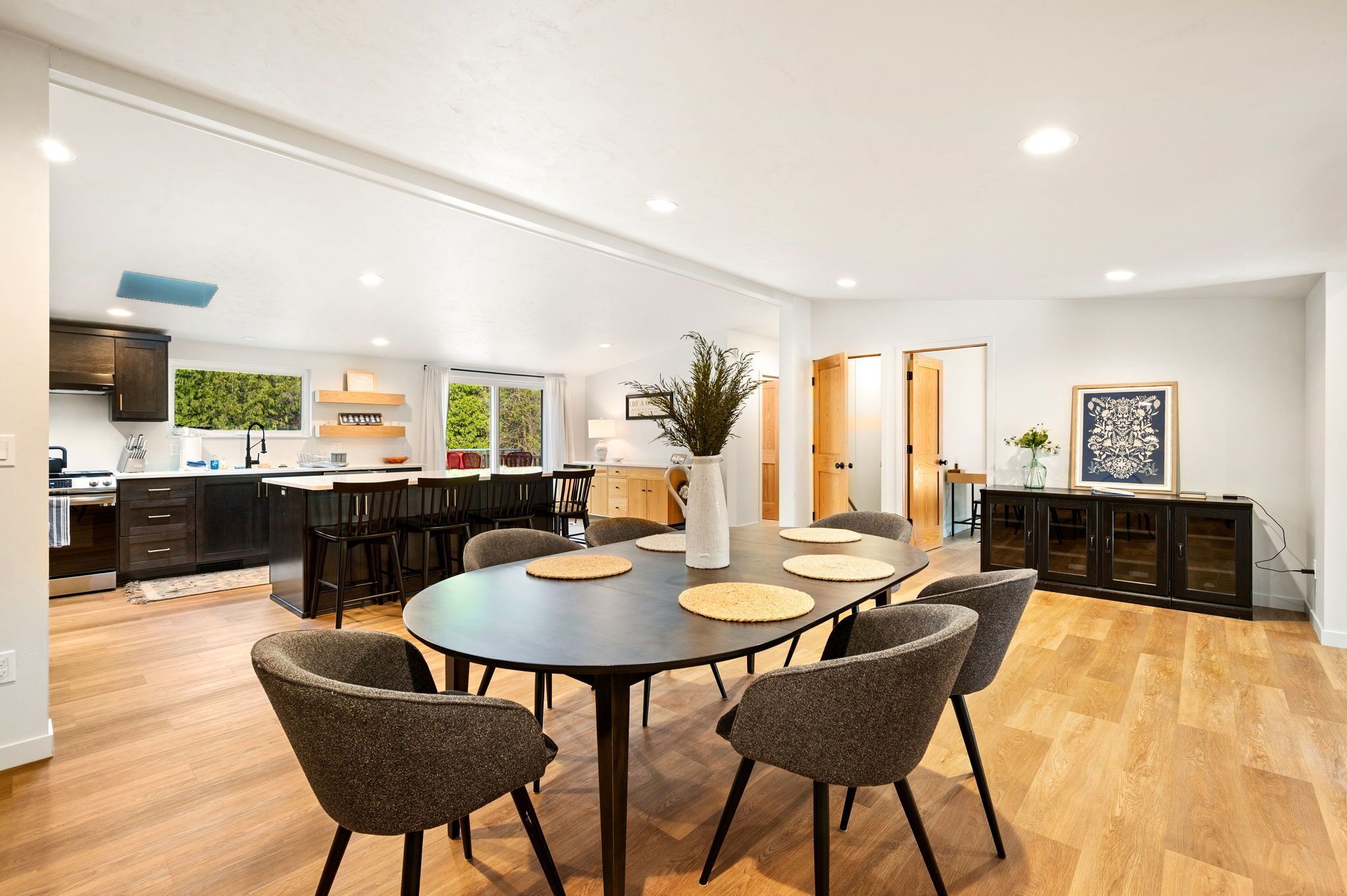 A modern kitchen with dark cabinets, a dining area with a round table and chairs, and a view of the outdoors through large windows.