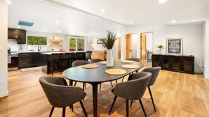 A modern kitchen with dark cabinets, a dining area with a round table and chairs, and a view of the outdoors through large windows.