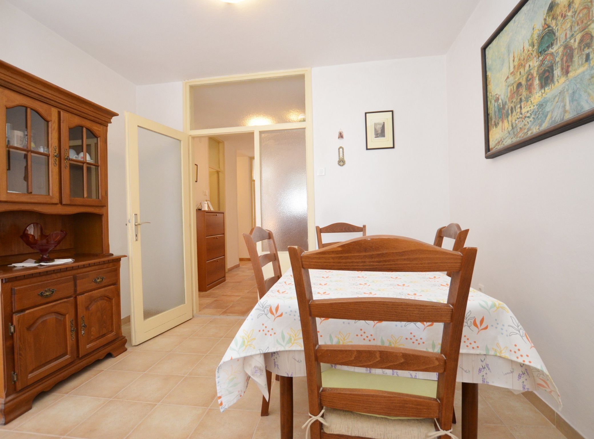 A room with a wooden dining table, chairs, a cabinet with glass doors, and a door leading to another room.