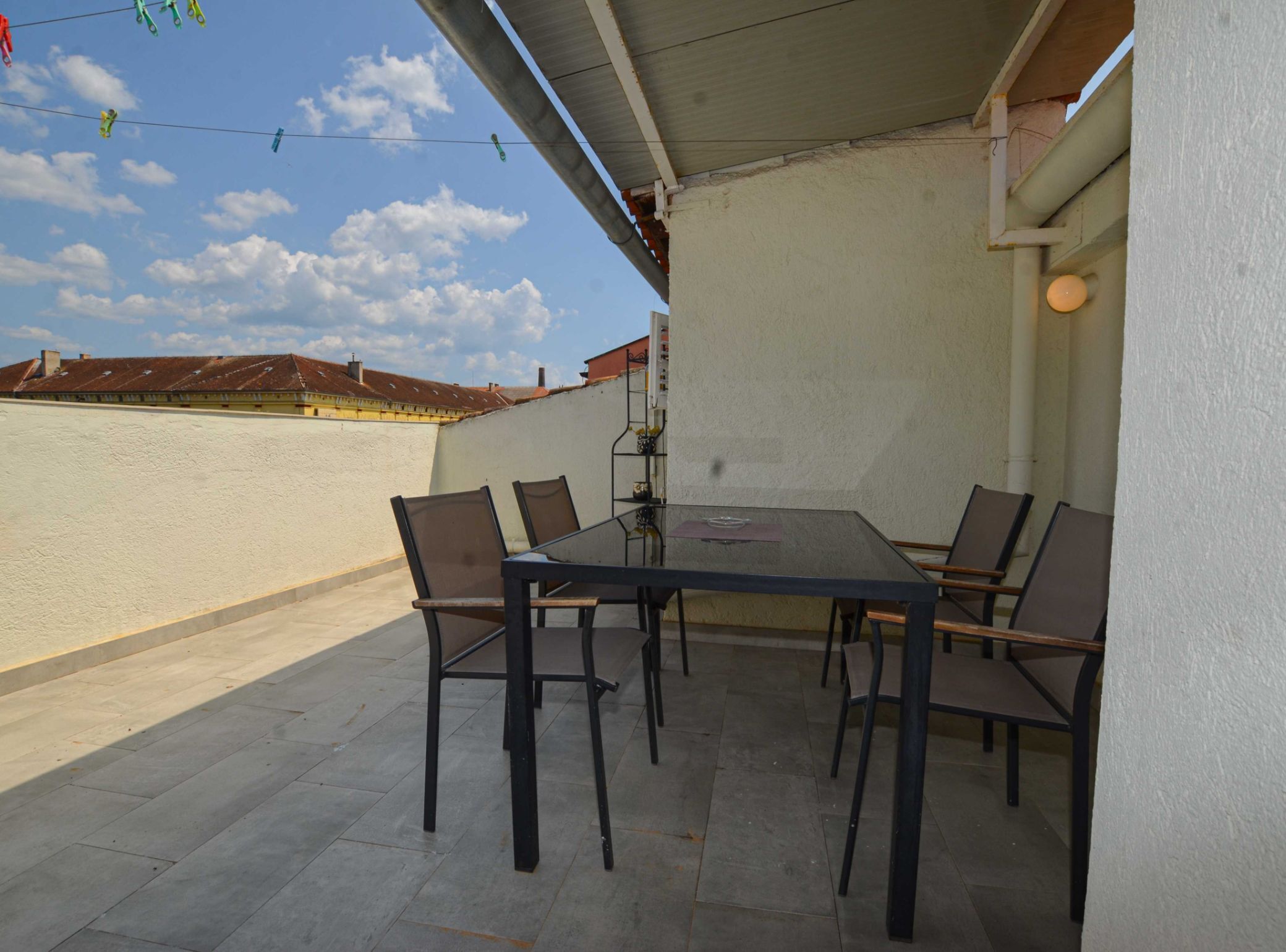 A covered outdoor patio with a table and chairs, a hanging plant, and a view of buildings in the background.