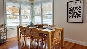 A dining area with a wooden table and wicker chairs, large windows with a view of a snowy street, and a framed artwork on the wall. - The Ingalls House - Downtown (Harbor Springs)