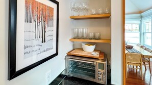 A kitchen with a framed artwork, a toaster oven, shelves with glasses, and a dining area with chairs. - The Ingalls House - Downtown (Harbor Springs)