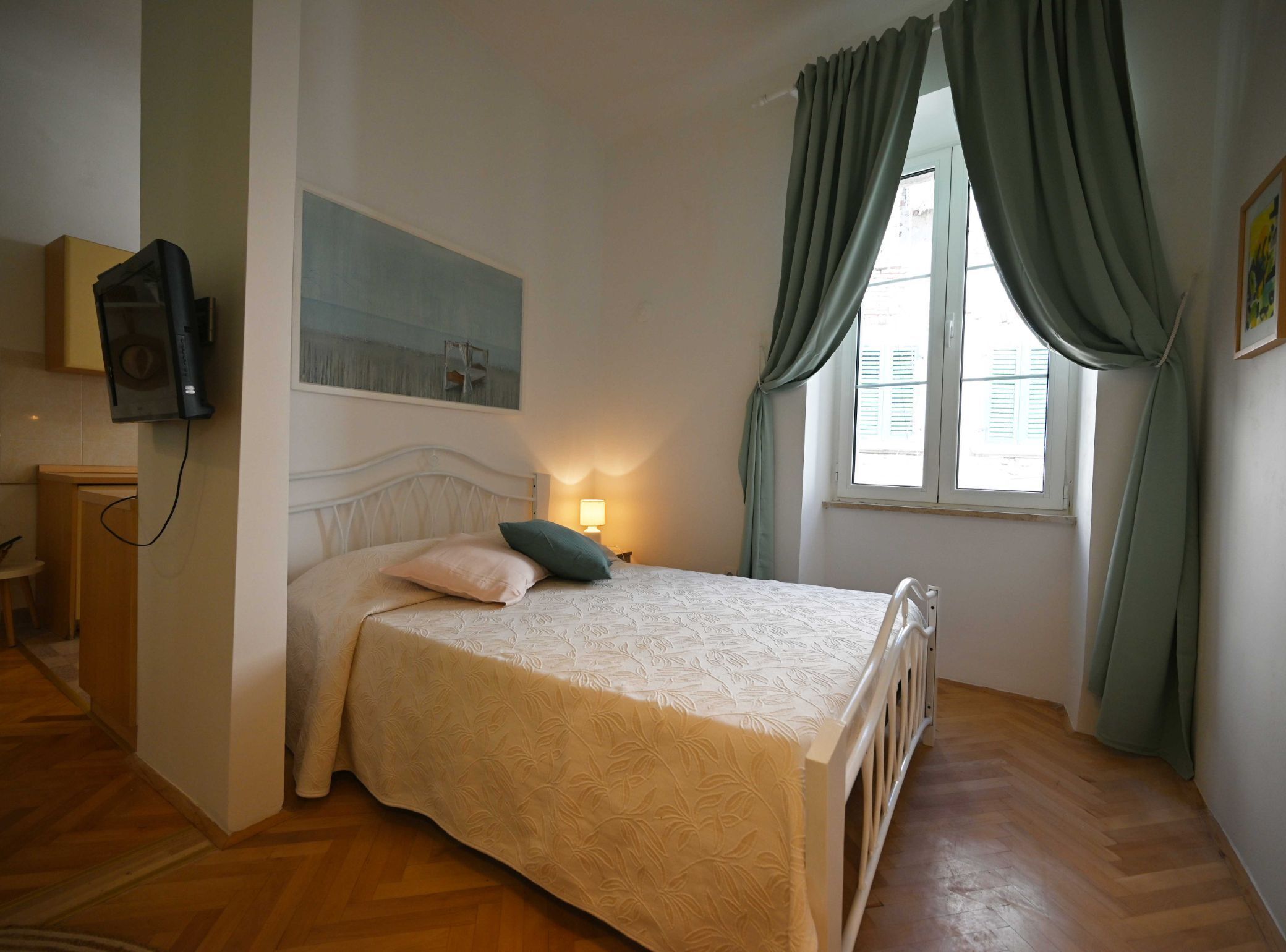 Een slaapkamer met een bed, een bedlampje, een raam met gordijnen, een aan de muur bevestigde televisie en een schilderij.