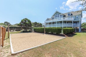 A sandy volleyball court with a wooden fence, a large two-story house with a balcony, and a well-maintained lawn.