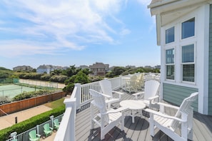 A deck with white chairs and a table, overlooking a tennis court and residential buildings. - Sun Realty gorgeous oceanside retreat with an elevator in Corolla NC! (Corolla)