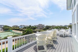 A wooden deck with white chairs and tables, overlooking a tennis court and residential buildings.