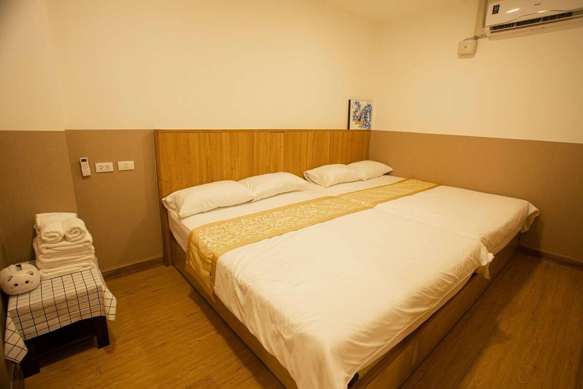 A hotel room with a wooden headboard, a bed with white bedding and a decorative gold runner, a small table with a checkered tablecloth and towels, and an air conditioning unit on the wall.