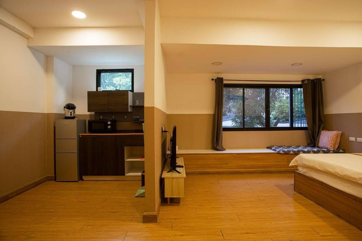 A compact room with a bed, a desk with a computer, a small kitchenette, and a window with curtains.