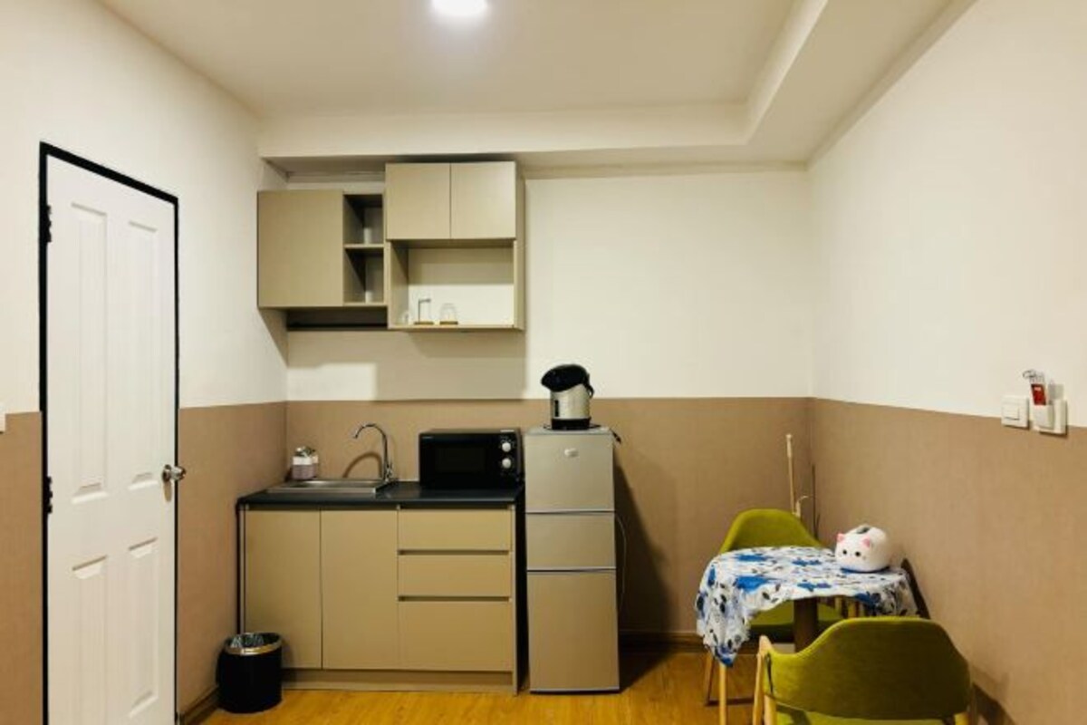 A compact kitchen with a microwave, kettle, and small refrigerator. There is a table with a tablecloth and a chair.