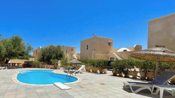 A pool area with lounge chairs and umbrellas, surrounded by buildings with a Mediterranean architectural style. - Sweet Villa Residence (Houmt Souk)