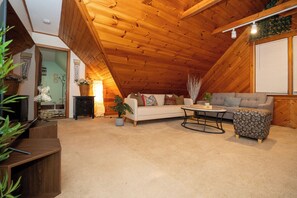 A cozy attic room with a wooden ceiling, a white sofa, a patterned ottoman, and a small table.