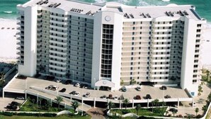 A high-rise hotel with multiple balconies, a central atrium, and a parking area with cars. - Admirals Quarter 908, 3 bedroom, 3 bath coastal oasis (Orange Beach)