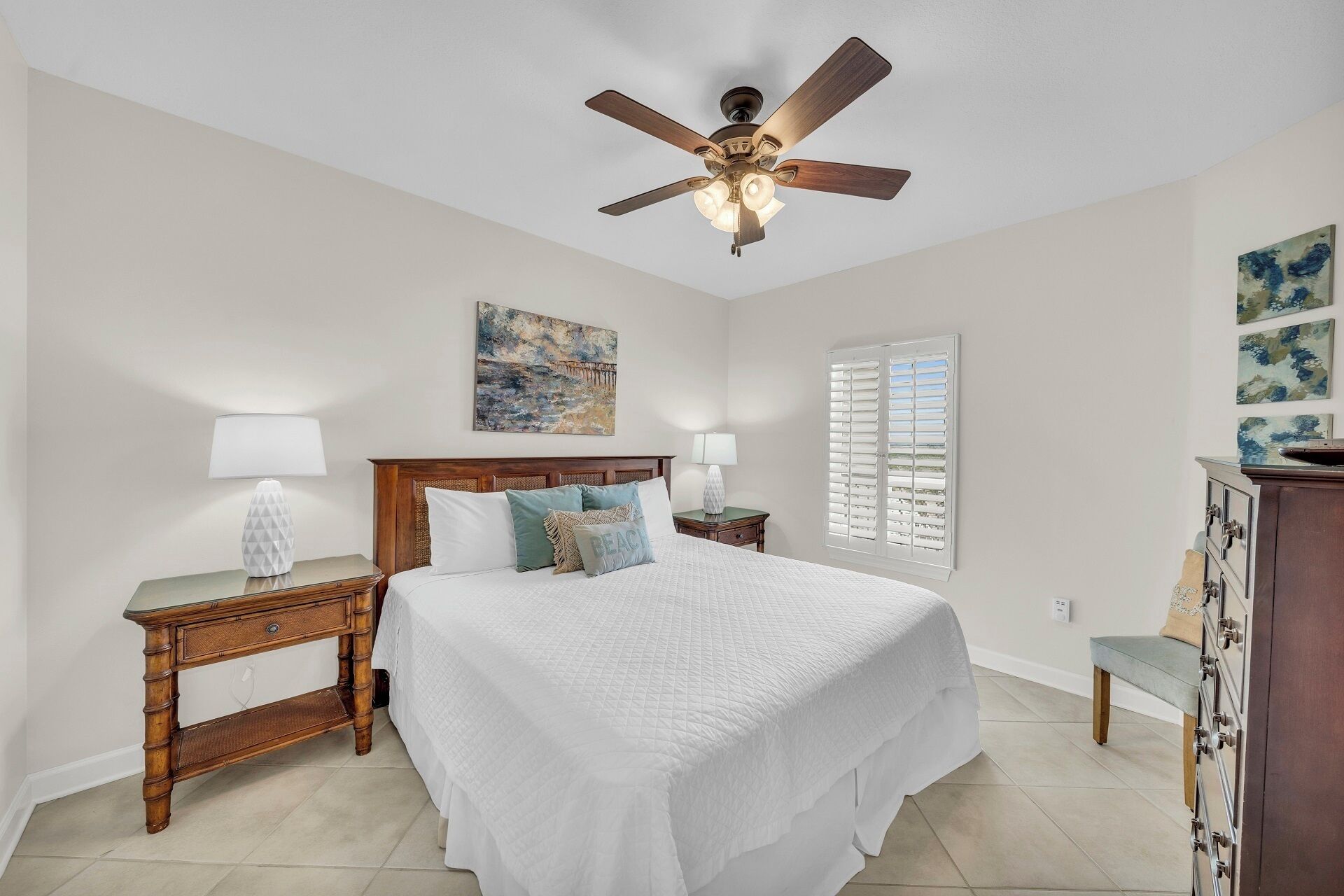 A bedroom with a bed, two bedside tables, a nightstand, a ceiling fan, a window with shutters, and artwork on the walls.