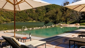Ein Poolbereich mit Liegestühlen und Sonnenschirmen, umgeben von einem See und bewaldeten Hügeln.